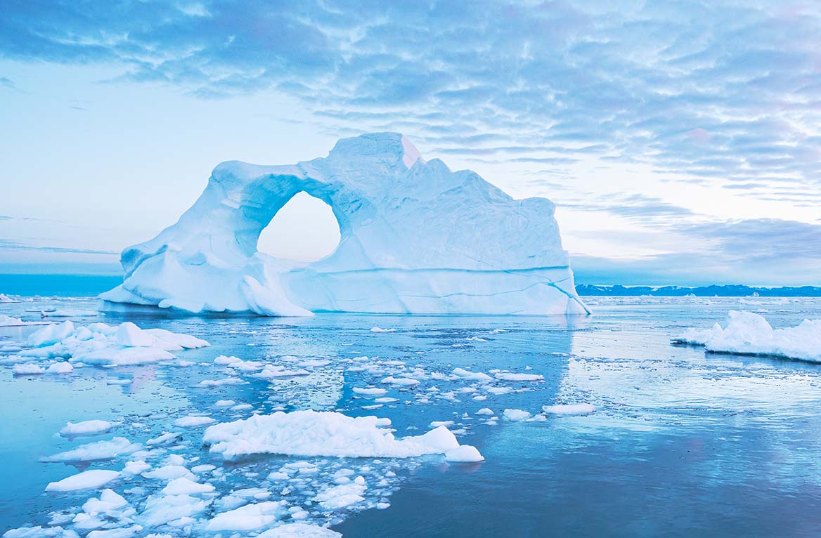 Sail boat with red sails cruising next to a pierced ice berg after sunset. Disko Bay, Greenland.